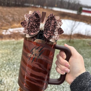May include: A brown and gold stainless steel travel mug with a black tiger stripe design and the word "STANLEY" printed on the side. The mug has a brown handle and a brown glitter bow with a gold glitter accent on top. The mug is sitting on a brown and gold beaded bracelet.