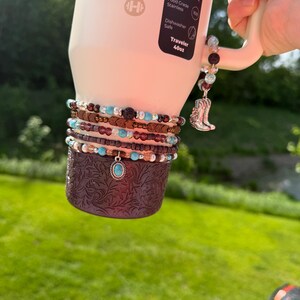 Puede incluir: Una botella de agua Traveler blanca de 1,18 litros con una funda marr&oacute;n estampada y m&uacute;ltiples pulseras de cuentas. Una pulsera tiene un colgante turquesa. Un dije con una bota de vaquero plateada cuelga del asa.