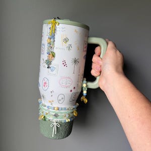 May include: A white travel mug with a green handle and base, decorated with olive branch and palm tree illustrations. The mug is adorned with beaded charms in shades of blue, yellow, and gold. The text "Olives" is visible on the mug.