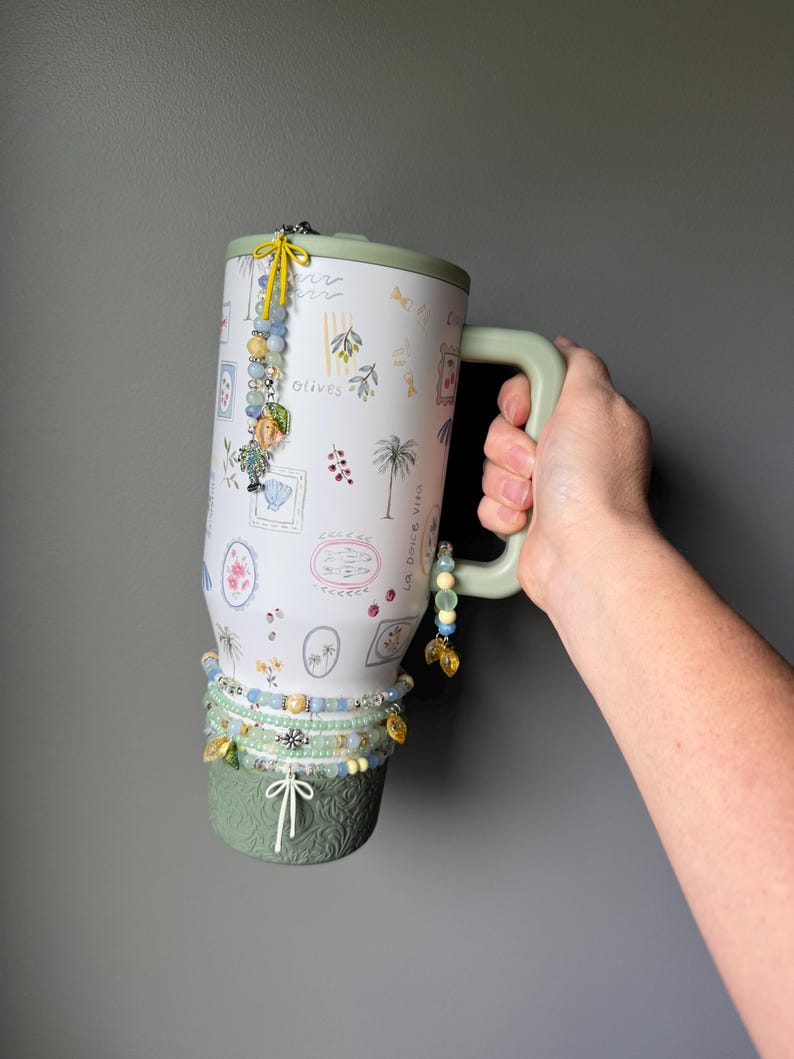 May include: A white travel mug with a green handle and base, decorated with olive branch and palm tree illustrations. The mug is adorned with beaded charms in shades of blue, yellow, and gold. The text "Olives" is visible on the mug.