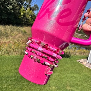 May include: A bright pink Stanley tumbler with the word "be" printed on it. Several pink and gold beaded bracelets are stacked on the tumbler. One bracelet has a pink heart charm. The tumbler is held outside on a sunny day.