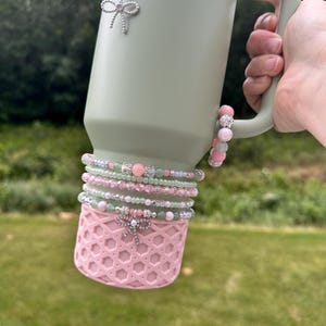 May include: A light green Stanley tumbler with a pink base and handle. The tumbler is adorned with beaded bracelets in pink, green, and white, and a silver bow charm. The word "STANLEY" is printed on the tumbler.