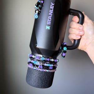 May include: Black Stanley mug with a handle, adorned with beaded bracelets in shades of purple, black, and blue. The mug features the Stanley logo in purple and is decorated with dangling beaded accents. The bottom of the mug has a decorative embossed pattern.