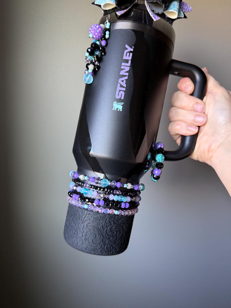 May include: Black Stanley mug with a handle, adorned with beaded bracelets in shades of purple, black, and blue. The mug features the Stanley logo in purple and is decorated with dangling beaded accents. The bottom of the mug has a decorative embossed pattern.
