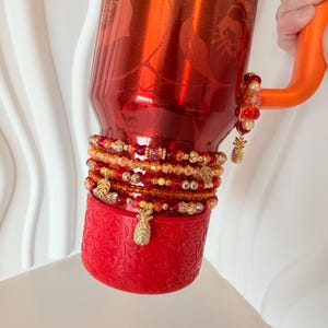 May include: A red insulated tumbler with a floral design and an orange handle. The tumbler is adorned with beaded bracelets in shades of red, gold, and orange, and gold pineapple charms. The base of the tumbler is red with a decorative pattern.