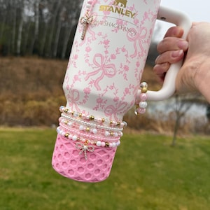 May include: A pink and white Stanley tumbler with a floral and bow pattern. The tumbler has a white handle and is adorned with beaded bracelets and a cross charm. The bottom of the tumbler is pink with a geometric design.