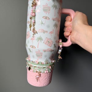 Puede incluir: Un vaso blanco con asa rosa que presenta un patr&oacute;n repetitivo de flamencos rosas, palmeras, coches antiguos, maletas y letreros de motel. El vaso est&aacute; decorado con un gran lazo brillante, abalorios de cuentas y una base rosa texturizada.