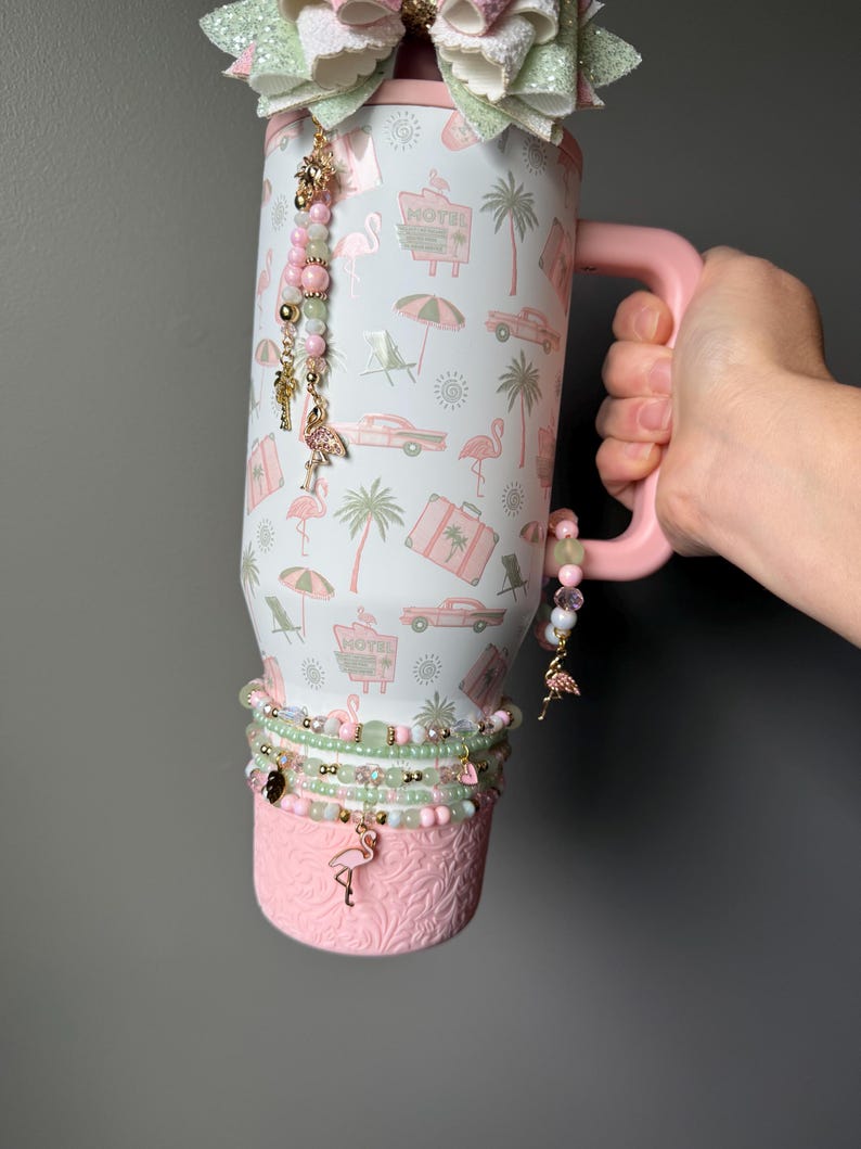 Puede incluir: Un vaso blanco con asa rosa que presenta un patr&oacute;n repetitivo de flamencos rosas, palmeras, coches antiguos, maletas y letreros de motel. El vaso est&aacute; decorado con un gran lazo brillante, abalorios de cuentas y una base rosa texturizada.
