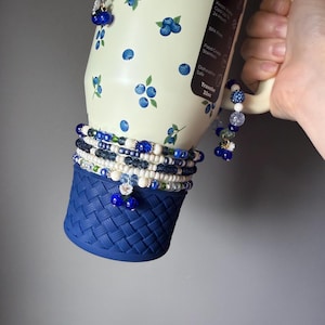 May include: A cream-colored 32oz tumbler with a blueberry pattern, featuring a dark blue woven-style silicone base boot. The tumbler is adorned with multiple beaded bracelets in shades of blue, white, and clear, featuring small flower and fruit charms.