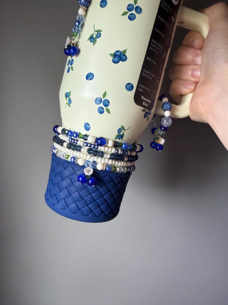 Op de afbeelding: Een cr&egrave;mekleurige 32oz beker met een bosbessenpatroon, voorzien van een donkerblauwe siliconen bodem met een geweven structuur. De beker is versierd met meerdere kralenarmbanden in blauwe, witte en transparante tinten met kleine bloem- en fruitbedels.