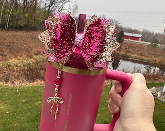 May include: A pink stainless steel tumbler with a gold lid and a pink and gold glitter bow. The tumbler has a gold handle and a gold charm with a pink ribbon. The tumbler is decorated with pink and gold beads. The tumbler says "Frappuccino + Starbucks" on the side.