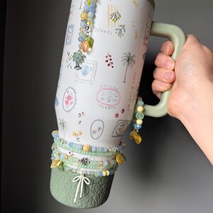 May include: A large, white travel mug with a green handle and base, decorated with olive branch and palm tree illustrations. The mug is adorned with beaded charms and bracelets in shades of blue, yellow, and green. The text "la dolce vita" is visible on the side.