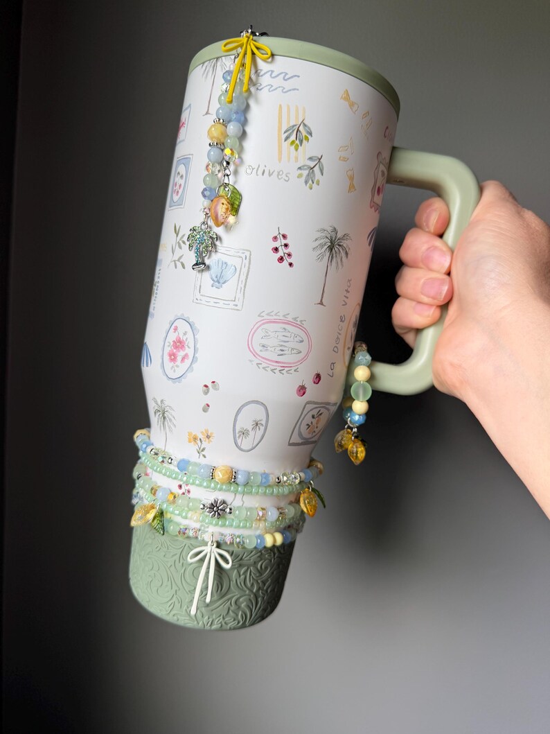 May include: A large, white travel mug with a green handle and base, decorated with olive branch and palm tree illustrations. The mug is adorned with beaded charms and bracelets in shades of blue, yellow, and green. The text "la dolce vita" is visible on the side.