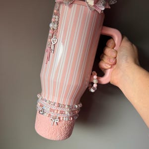 Pu&ograve; includere: Un bicchiere a righe rosa chiaro e bianco con manico, sormontato da un grande fiocco glitterato rosa e argento. Il bicchiere presenta una base testurizzata, un ciondolo di perline con una chiave a forma di cuore e braccialetti di perline.
