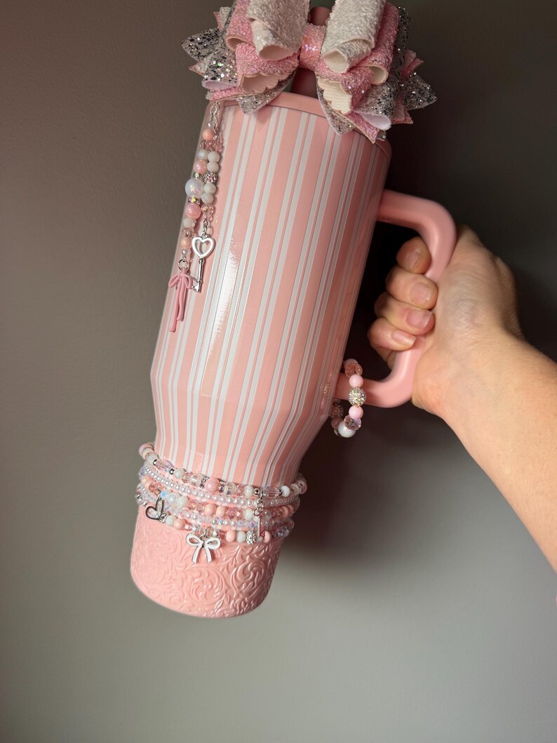 Pu&ograve; includere: Un bicchiere a righe rosa chiaro e bianco con manico, sormontato da un grande fiocco glitterato rosa e argento. Il bicchiere presenta una base testurizzata, un ciondolo di perline con una chiave a forma di cuore e braccialetti di perline.