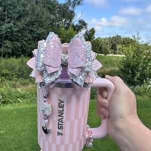 May include: A pink Stanley mug with a decorative bow, beaded charms, and a handle. The mug has a vertical striped pattern and is embellished with rhinestones and beads. The bow is pink and silver glitter.