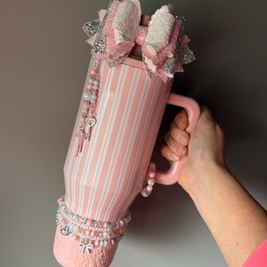 Pu&ograve; includere: Un bicchiere rosa chiaro con strisce verticali bianche, caratterizzato da un grande fiocco glitterato a strati rosa e argento sulla parte superiore. Il bicchiere &egrave; decorato con ciondoli di perline pendenti, tra cui una chiave e un fiocco.