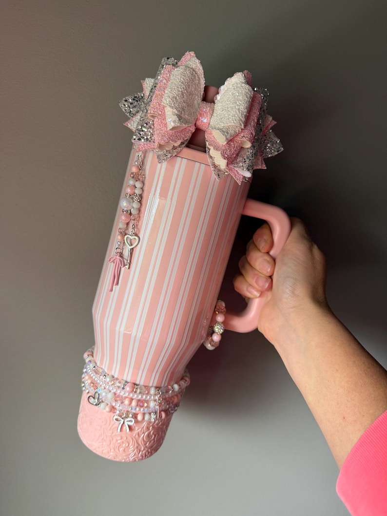 Pu&ograve; includere: Un bicchiere rosa chiaro con strisce verticali bianche, caratterizzato da un grande fiocco glitterato a strati rosa e argento sulla parte superiore. Il bicchiere &egrave; decorato con ciondoli di perline pendenti, tra cui una chiave e un fiocco.