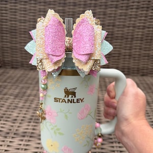 May include: A light green Stanley tumbler with a floral design, held by a hand. The tumbler is adorned with a decorative bow in pink, gold, and mint green. The bow is on top of the tumbler, and the handle is on the right side. The tumbler has a straw.
