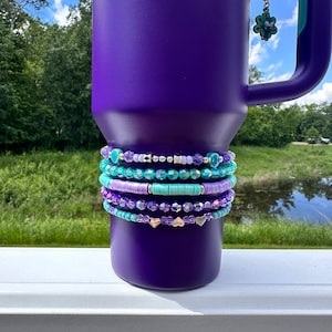 May include: A purple tumbler with a turquoise and purple beaded charm. The charm is made of small beads and has a heart-shaped charm.