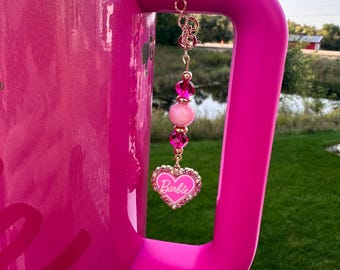 May include: Pink travel mug with a gold chain charm with pink and clear beads and a pink heart charm with the word "Barbie" on it.