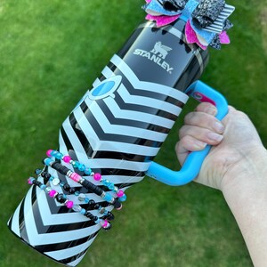 May include: A black and white striped stainless steel water bottle with a blue handle and a black, white and pink bow on the lid. The bottle has the word "STANLEY" printed on the side. The bottle is surrounded by a stack of colourful beaded bracelets.