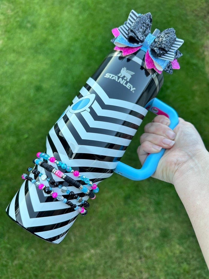 May include: A black and white striped stainless steel water bottle with a blue handle and a black, white and pink bow on the lid. The bottle has the word "STANLEY" printed on the side. The bottle is surrounded by a stack of colourful beaded bracelets.
