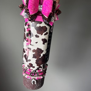 May include: A stainless steel tumbler featuring a brown and white cow print pattern, topped with a large, multi-layered glittery pink and brown bow. The tumbler is adorned with beaded charms, including a pink cowboy boot and a horseshoe charm.