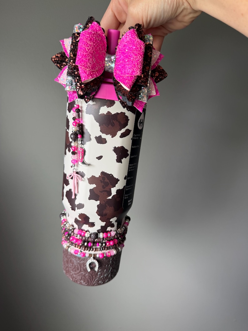 May include: A stainless steel tumbler featuring a brown and white cow print pattern, topped with a large, multi-layered glittery pink and brown bow. The tumbler is adorned with beaded charms, including a pink cowboy boot and a horseshoe charm.
