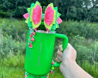 Pode incluir: Um copo verde brilhante com uma alça, adornado com um laço com tema de melancia e detalhes de contas. O copo apresenta um design rosa e verde, com uma base rosa e um pingente de melancia. O copo é segurado por uma mão.