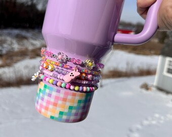 Peut inclure: Un gobelet isotherme violet clair avec une poignée, orné d'une pile de bracelets de perles colorés. Les bracelets comportent diverses perles, dont des fleurs, des visages souriants et un charm de lapin rose. Le bas du gobelet présente un motif à carreaux pastel.