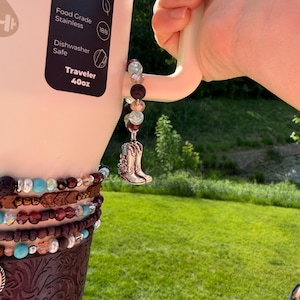 Puede incluir: Una botella de agua Traveler blanca de 1,18L con un charm decorativo. El charm presenta una bota de vaquero plateada, cuentas y una envoltura de cuero marr&oacute;n. La botella es sostenida por una mano, con un fondo de hierba verde.