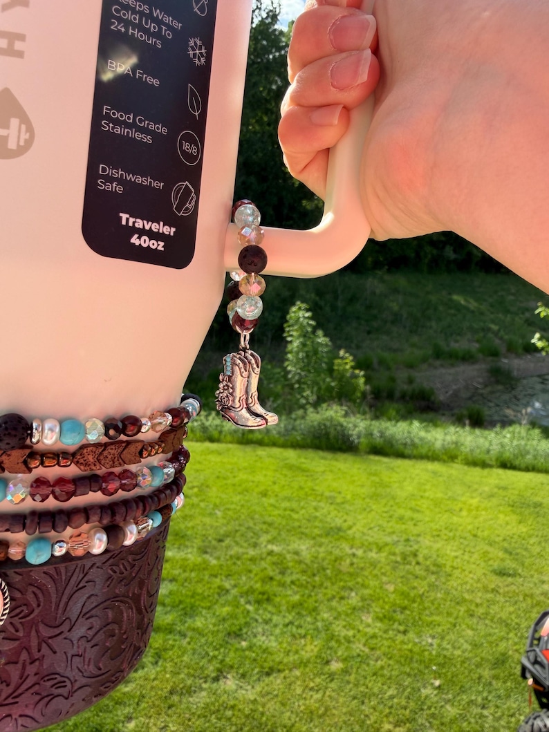 Puede incluir: Una botella de agua Traveler blanca de 1,18L con un charm decorativo. El charm presenta una bota de vaquero plateada, cuentas y una envoltura de cuero marr&oacute;n. La botella es sostenida por una mano, con un fondo de hierba verde.