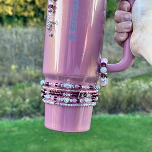 Op de afbeelding: Een roze Stanley-beker met bijpassende handgreep, versierd met kralenarmbanden en bedels. De armbanden hebben roze, zilveren en bordeauxrode kralen, met hartvormige accenten. Een sleutelhanger en een strass-hart hangen aan de beker.