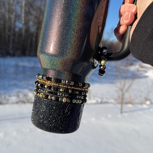 May include: A black and iridescent tumbler with a handle, adorned with beaded bracelets. The tumbler has a textured base and the word "STAN" in gold lettering. The background is a snowy landscape under a blue sky.