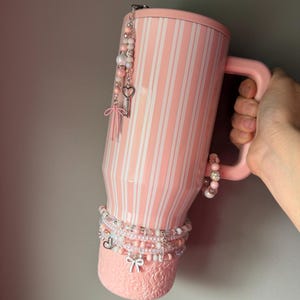 Pu&ograve; includere: Un bicchiere a righe rosa chiaro e bianco con manico, caratterizzato da una base testurizzata con motivo floreale. Il bicchiere &egrave; decorato con diversi ciondoli di perline, tra cui una chiave argentata a forma di cuore e un piccolo fiocco.