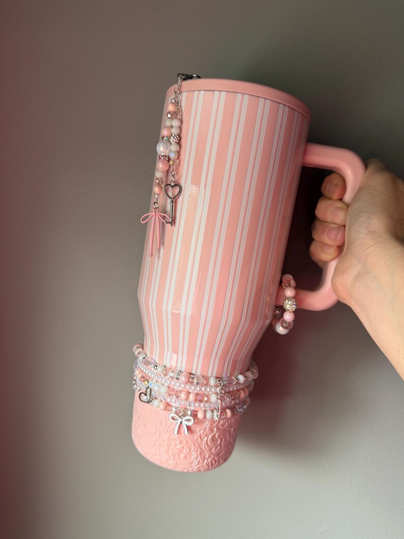 Pu&ograve; includere: Un bicchiere a righe rosa chiaro e bianco con manico, caratterizzato da una base testurizzata con motivo floreale. Il bicchiere &egrave; decorato con diversi ciondoli di perline, tra cui una chiave argentata a forma di cuore e un piccolo fiocco.