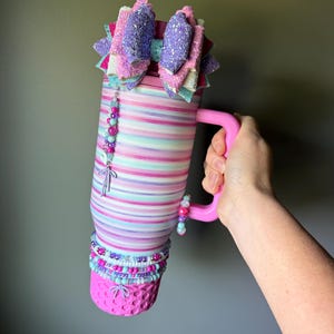 Op de afbeelding: Een hoge, cilindrische beker met een roze handvat, voorzien van horizontale strepen in tinten roze, paars en lichtblauw. De bovenkant is versierd met een grote, meerlaagse glitterstrik en het lichaam is gedecoreerd met kralenbedels en armbanden.