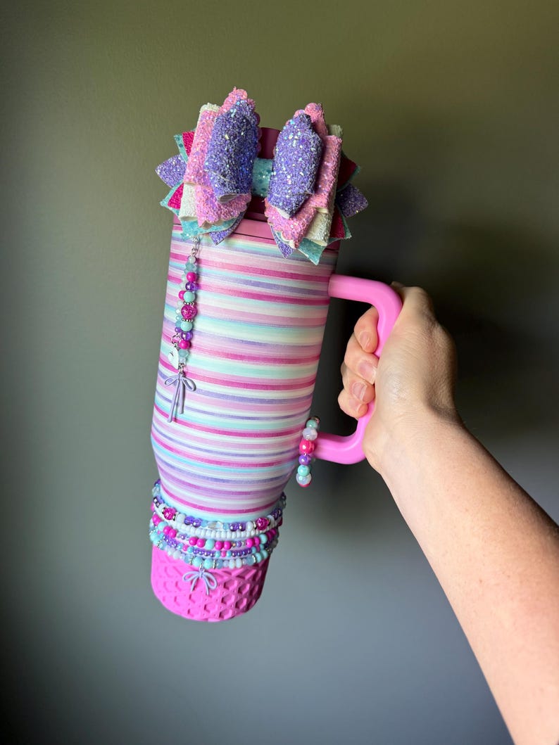 Op de afbeelding: Een hoge, cilindrische beker met een roze handvat, voorzien van horizontale strepen in tinten roze, paars en lichtblauw. De bovenkant is versierd met een grote, meerlaagse glitterstrik en het lichaam is gedecoreerd met kralenbedels en armbanden.