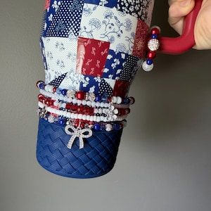 Puede incluir: Un vaso grande con un patr&oacute;n floral de retazos en rojo, blanco y azul y una base texturizada de color azul s&oacute;lido. El vaso cuenta con un asa roja adornada con un dije de cuentas y varias pulseras de cuentas apiladas con dijes de lazo y flor.