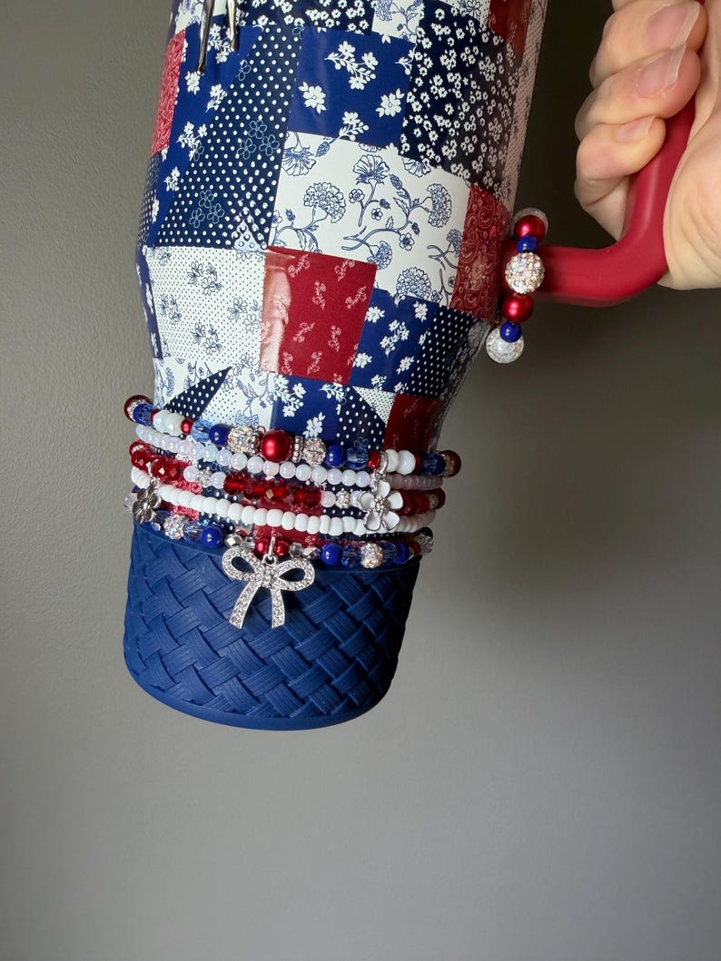 Puede incluir: Un vaso grande con un patr&oacute;n floral de retazos en rojo, blanco y azul y una base texturizada de color azul s&oacute;lido. El vaso cuenta con un asa roja adornada con un dije de cuentas y varias pulseras de cuentas apiladas con dijes de lazo y flor.