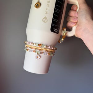 May include: A cream-colored 40oz HydroJug tumbler with a handle, featuring a vertical black label with white text. The tumbler is adorned with several beaded accessories, including a sunflower charm and a white daisy charm on beaded strands.
