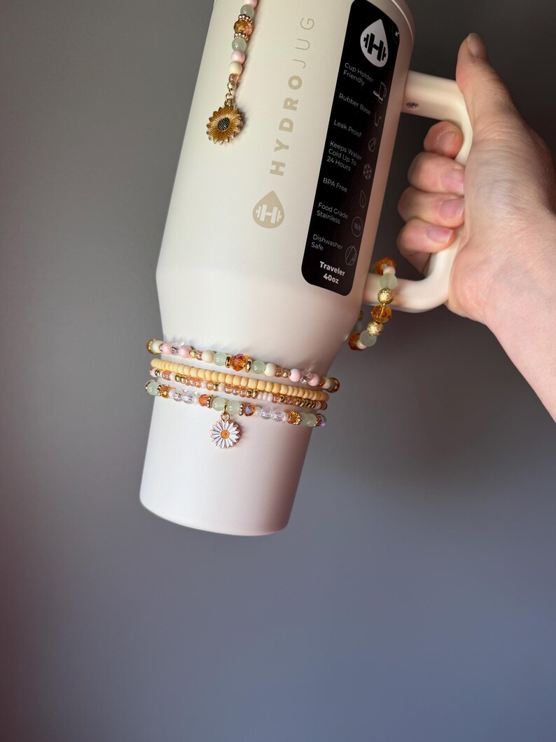 May include: A cream-colored 40oz HydroJug tumbler with a handle, featuring a vertical black label with white text. The tumbler is adorned with several beaded accessories, including a sunflower charm and a white daisy charm on beaded strands.