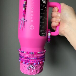 May include: A bright pink HydroJug water bottle with a handle, adorned with beaded bracelets and charms in shades of pink and purple. The bottle has the text "HYDROJUG" and "Traveler 32oz" printed on it.