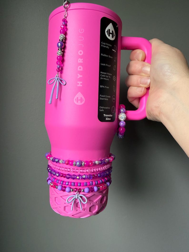 May include: A bright pink HydroJug water bottle with a handle, adorned with beaded bracelets and charms in shades of pink and purple. The bottle has the text "HYDROJUG" and "Traveler 32oz" printed on it.