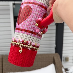 May include: A red handled tumbler with a mosaic heart design in pink, white, and gold. The tumbler has a red knitted sleeve and is adorned with beaded bracelets in shades of red, pink, and gold. A gold heart-shaped charm hangs from the bracelets.