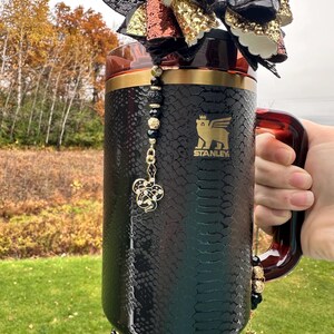 Peut inclure: Mug Stanley noir avec un motif peau de serpent, des détails dorés et un charm décoratif. Le mug a une anse et est orné d'un charm perlé et d'un nœud décoratif. Le logo Stanley est visible sur le côté.