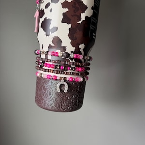 May include: A stainless steel tumbler with a brown and white cow print pattern, featuring a stack of beaded bracelets wrapped around its base. The bracelets include pink, brown, and silver beads with a silver horseshoe charm hanging from the center.