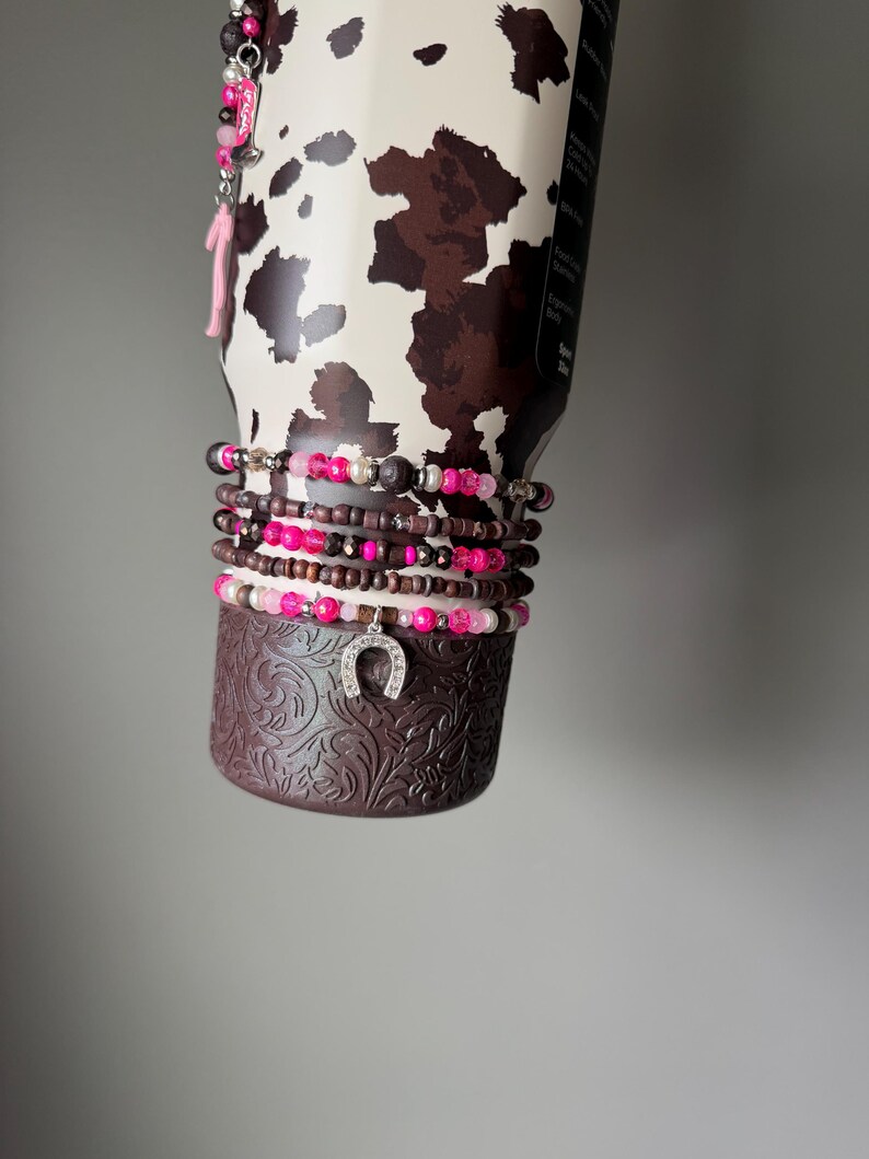 May include: A stainless steel tumbler with a brown and white cow print pattern, featuring a stack of beaded bracelets wrapped around its base. The bracelets include pink, brown, and silver beads with a silver horseshoe charm hanging from the center.