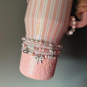Pu&ograve; includere: Un bicchiere rosa chiaro con strisce verticali bianche e una base floreale testurizzata. Il bicchiere &egrave; decorato con una pila di braccialetti di perline con perle rosa e bianche, cristalli trasparenti e ciondoli argentati a forma di fiocco, cuore e chiave.